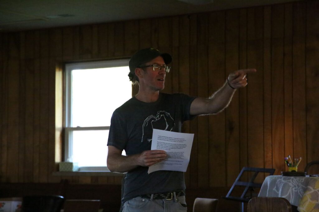 A man wearing a baseball cap and holding a paper gestures with his hand.